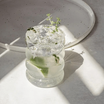 Ein mit Wasser, Limone, Eiswürfeln und Kräutern befülltes Bubble Glas Blanko von Eulenschnitt steht neben einem Tablett. North Glow