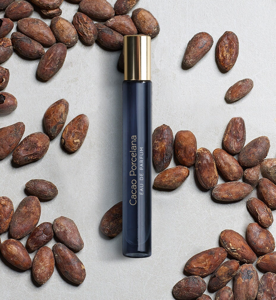 Dunkelblaues schmales Glasgefäß mit goldenem Deckel "Cacao Porcelana" von Atelier Materi umgeben von Kakaobohnen.