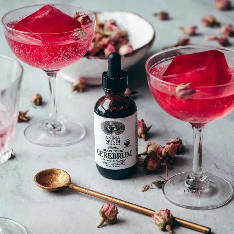 Anima Mundi Cerebrum Pipettenglasflasche steht zwischen 2 Gläsern mit roter Flüssigkeit, davor liegt ein Messinglöffel.