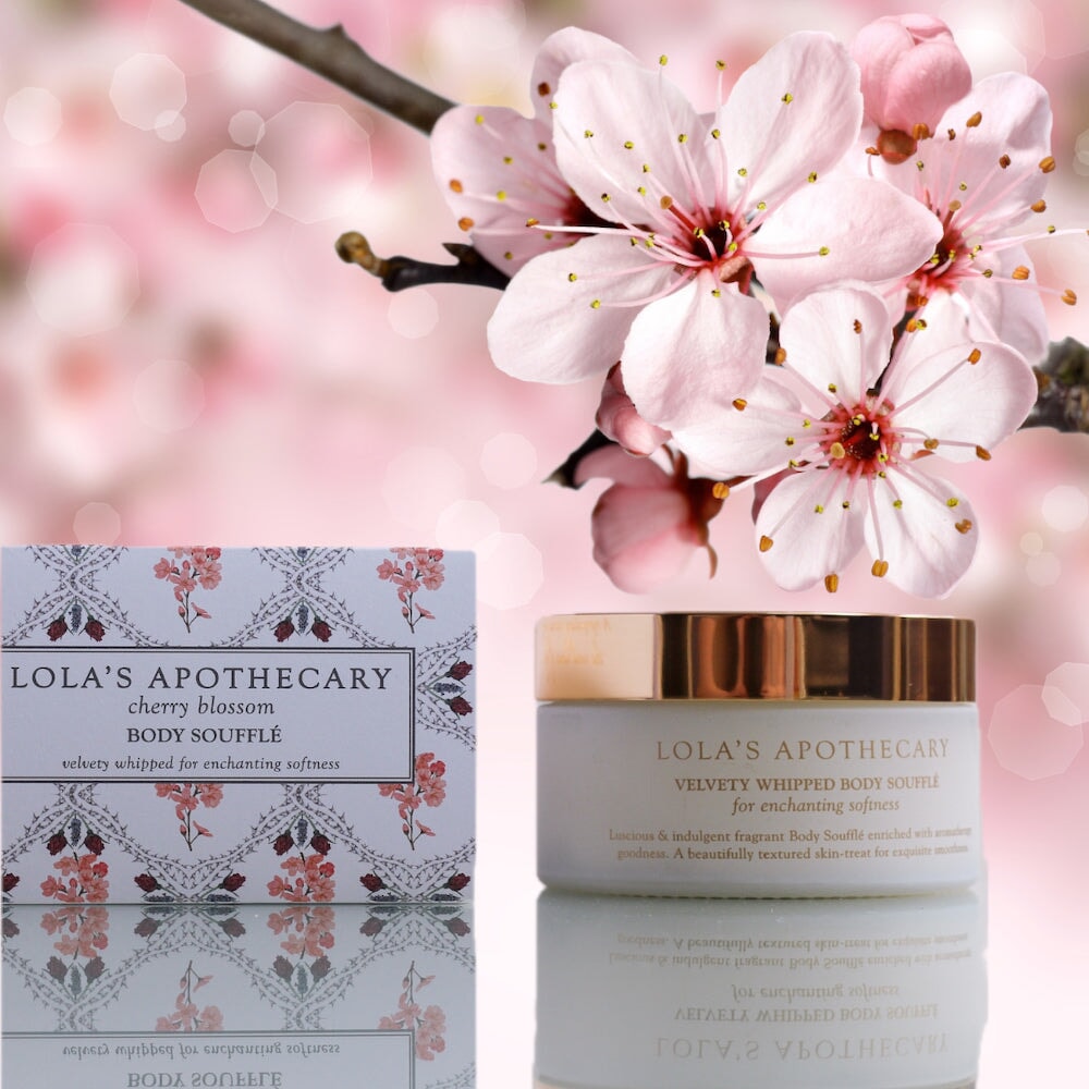Weißer Tiegel  mit goldenem Decke Cherry Blossom Body Soufflé von Lola's Apothecary steht neben der Verpackung vor Kirschblüten.