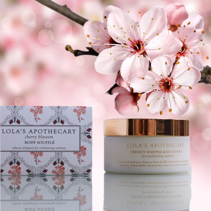 Weißer Tiegel  mit goldenem Decke Cherry Blossom Body Soufflé von Lola's Apothecary steht neben der Verpackung vor Kirschblüten.