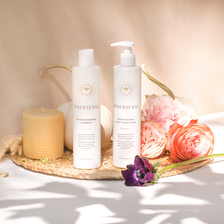 Innersense Color Radiance Daily Conditioner und Color Awakening Shampoo Flaschen stehen auf einer Bastmatte mit Blumendekoration und Bienenwachskerze. 