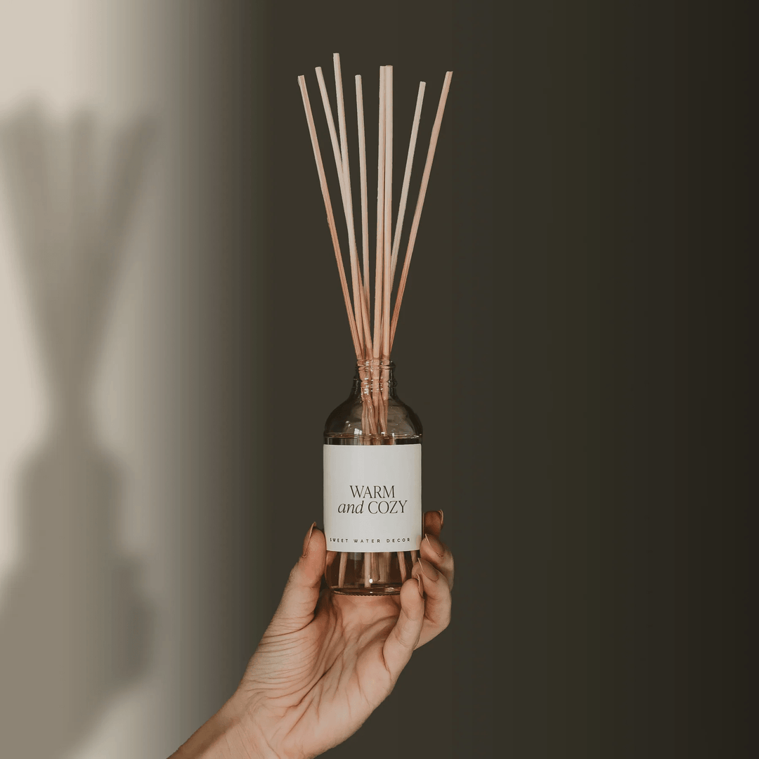 Hand hält Sweet Water Decor Diffuser Warm and Cozy Flasche mit Schilfrohtstäbchen. North Glow