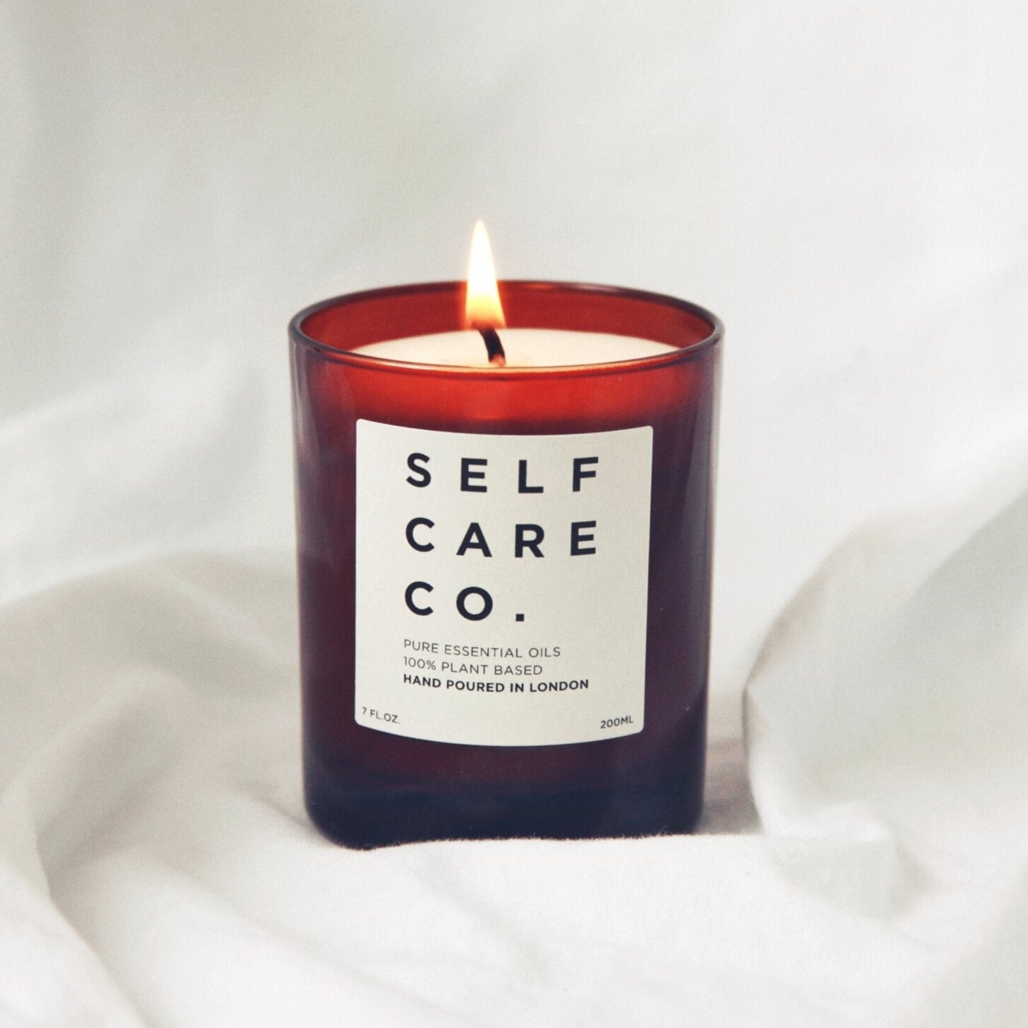 Braunes Kerzenglas mit der Aufschrift Self Care Co. steht auf einem weißen Tuch
