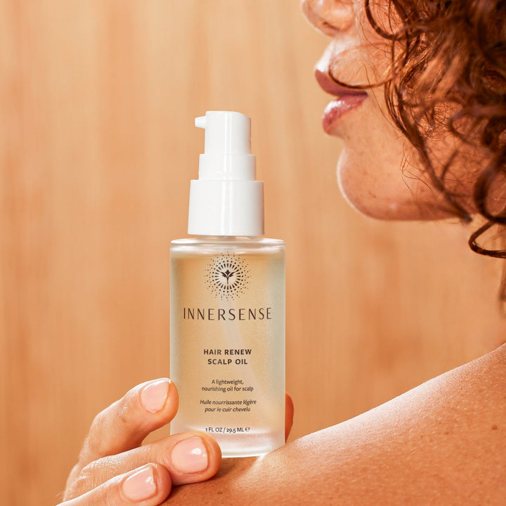 Ausschnitt eines Frauenkopfes mit Schulter in warmen Farben, Hand hält Innersense Hair Renew Scalp Oil Flasche auf der Schulter fest.