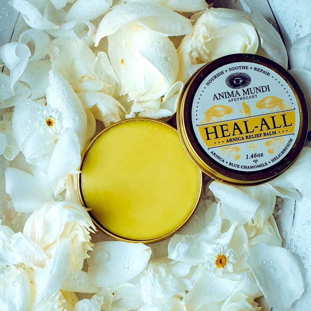 Offene Anima Mundi Heal-All Balm Dose mit gelbem Inhalt, der Deckel liegt daneben zwischen weißen Blütenblättern.