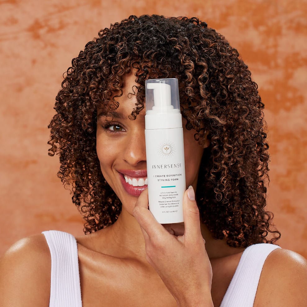 INNERSENSE I Create Definition Pumpflasche gehalten von einer Frauenhand vor einem Frauenkopf mit stark gelockten Haaren.