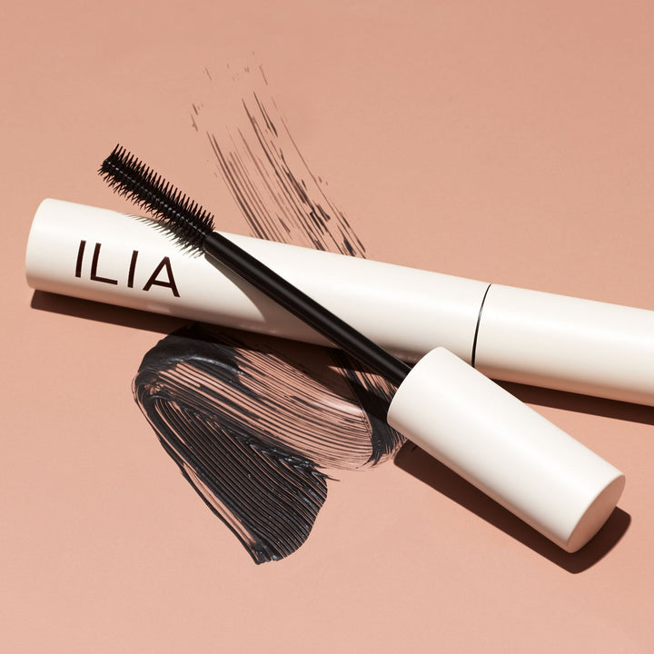 extur- und Farbbeispiel der Ilia Limitless Lash Mascara mit  offener Mascara und danebenliegender Bürste vor hellbraunem Hintergrund.
