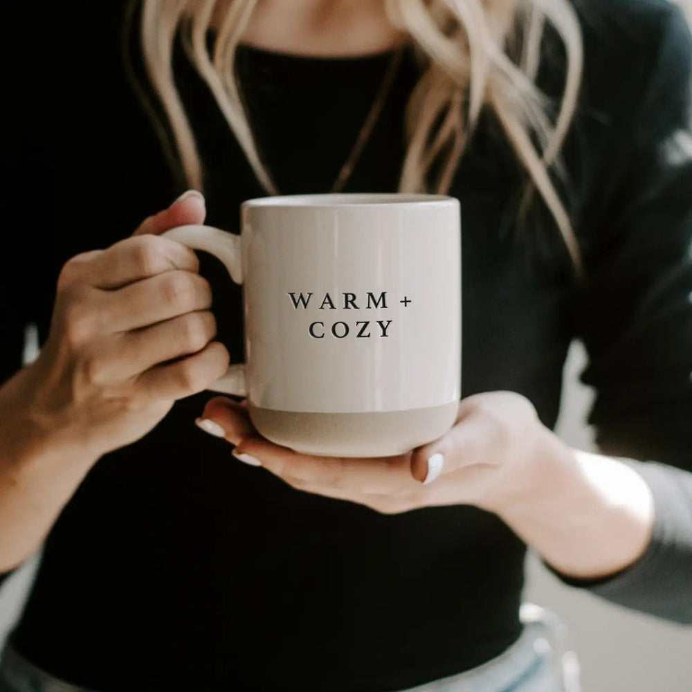 Person hält Warm and Cozy Tasse mit beiden Händen, kuscheliges Herbst-Outfit im Hintergrund.