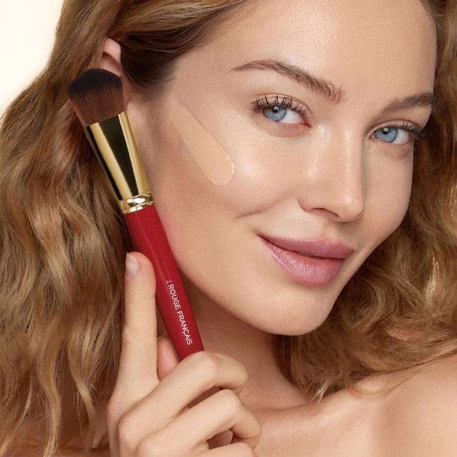 Nahaufnahme eines Frauengesichts mit einem Streifen flüssiger Foundation auf der Wange, in der Hand hält sie den Le Rouge Francais Liquid Foundation Brush No 810.