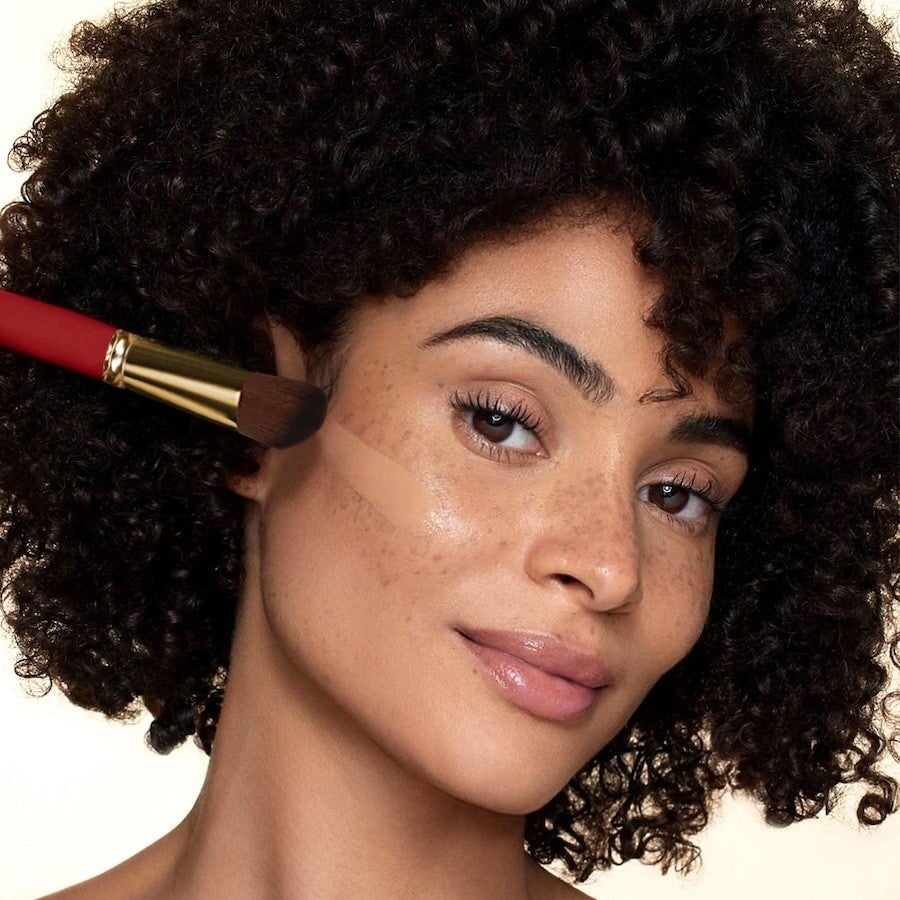 Nahaufnahme eines Frauengesichts mit dunklen Locken mit einem Streifen flüssiger Foundation auf der Wange, in der Hand hält sie den Le Rouge Francais Liquid Foundation Brush No 810. North Glow