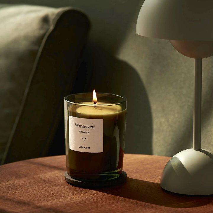 Looops Kerze Winterzeit in grünem Glas steht auf einem hölzernen Beistelltisch neben einer Lampe.