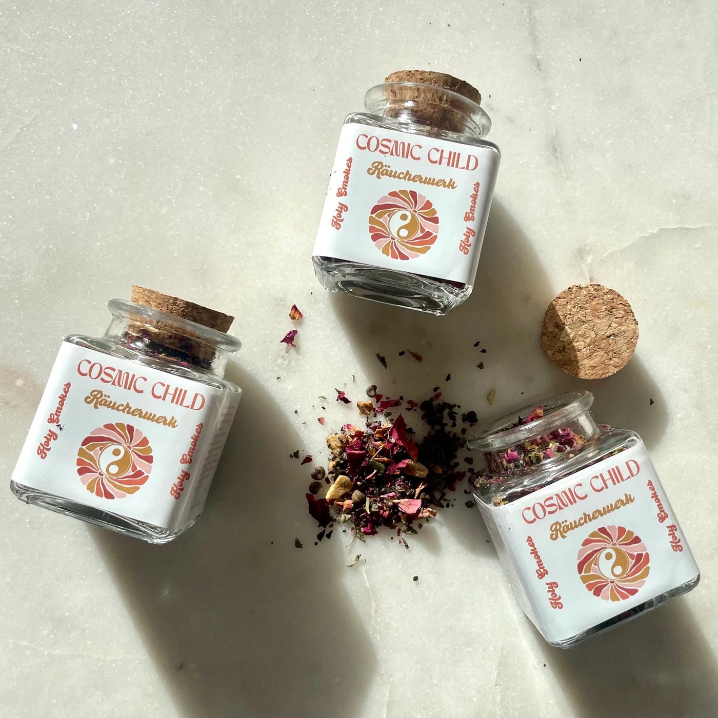 3 quadratische Glasflaschen Cosmic Child Räucherwerk mit Korkverschluss, eine davon geöffnet und getrocknete Blüten und Blätter liegen verstreut dazwischen.