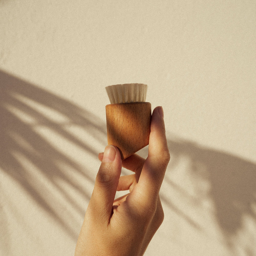 Eine Hand hält die ruhi Vegan Facial Dry Brush vor einer hellen Wand mit ästhetischem Palmenschatten.