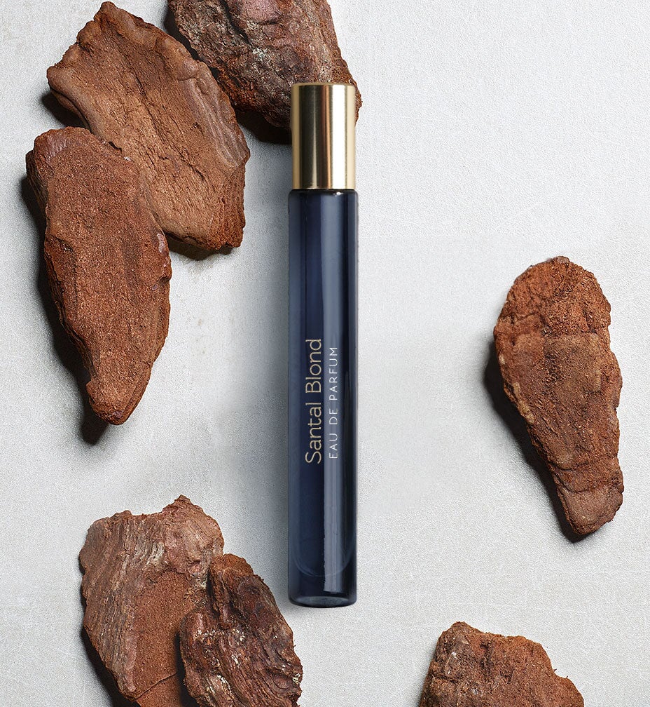 Dunkelblaues, schmales Glasgefäß mit goldenem Deckel  "Santal Blond" von Atelier Materi umgeben von kleinen Holzstücken.