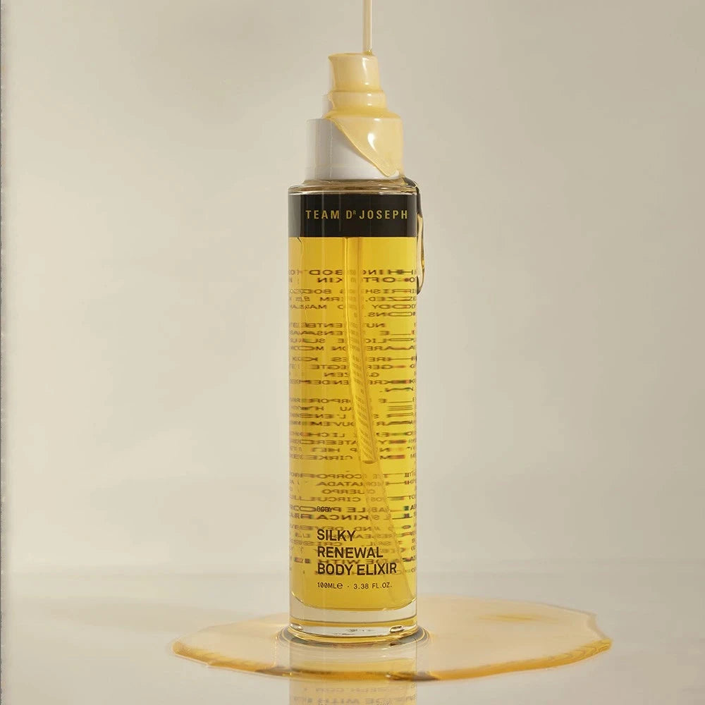 Team Dr Joseph Silky Renewal Body Elixir – transparente Glasflasche mit goldgelbem Körperöl und Pumpspender, übergossen mit Öl, das am Flaschenhals hinabläuft und sich dekorativ auf der hellen Oberfläche verteilt.