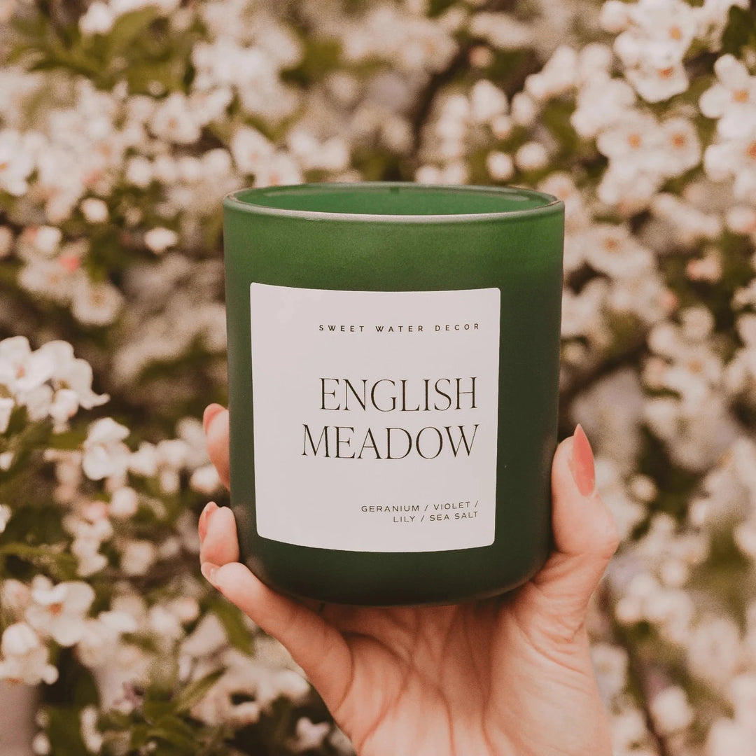 Duftkerze in grünem Milchglas English Meadow von Swee Water Decor, gehalten von einer Hand, im Hintergrund verschwommen Buschblumen.
