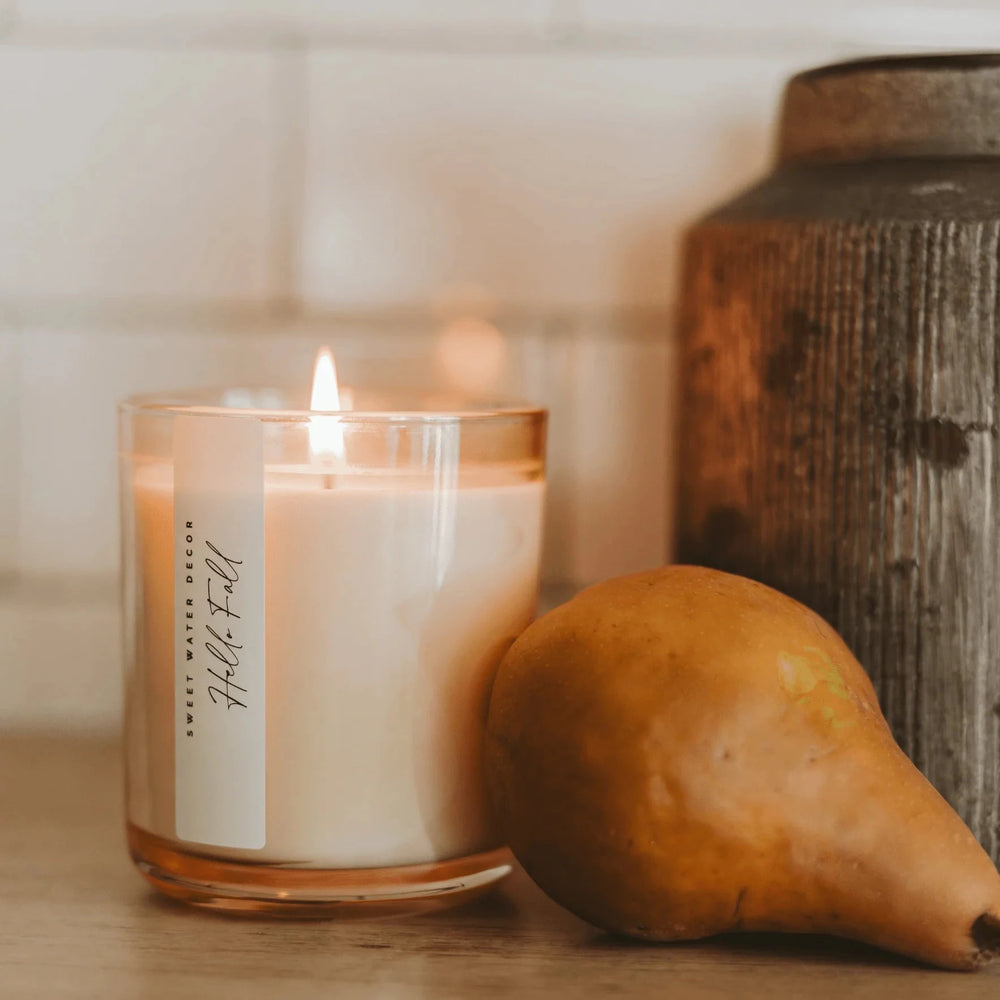 Brennende Hello Fall Sojawachskerze von Sweet Water Decor im beige-getöntem Glas platziert auf einem Tisch neben einer reifen Birne