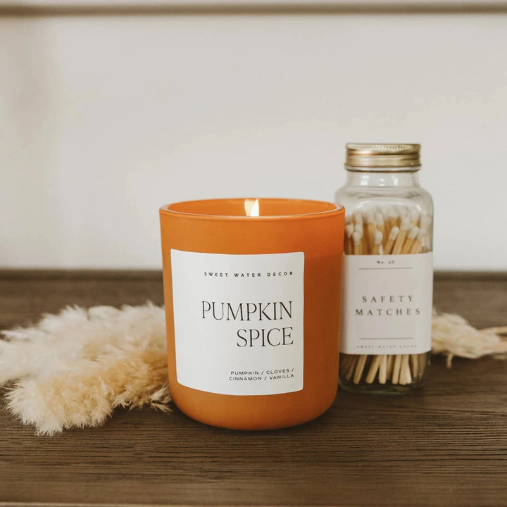 Brennende Pumpkin Spice Sojawachskerze von Sweet Water Decor im orangefarbenen Mattglas auf einem Holztisch neben einem Schraubglas Streichhölzer