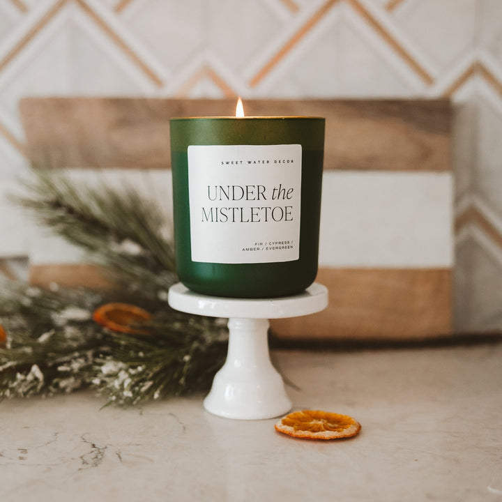 Duftkerze Under the Mistletoe von Sweet Water Decor steht auf einem kleinem Podest im Vordergrund, im Hintergrund sind Tannenzweige.