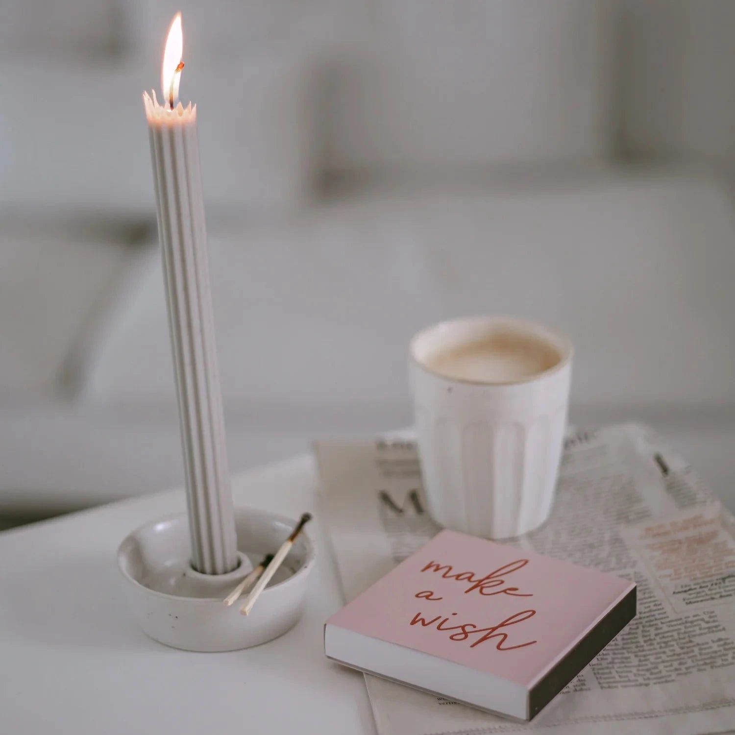 Streichholzschachtel mit Aufschrift "make a wish", liegend auf einer Zeitung, daneben eine brennende Stabkerze und ein Becher Kaffee.