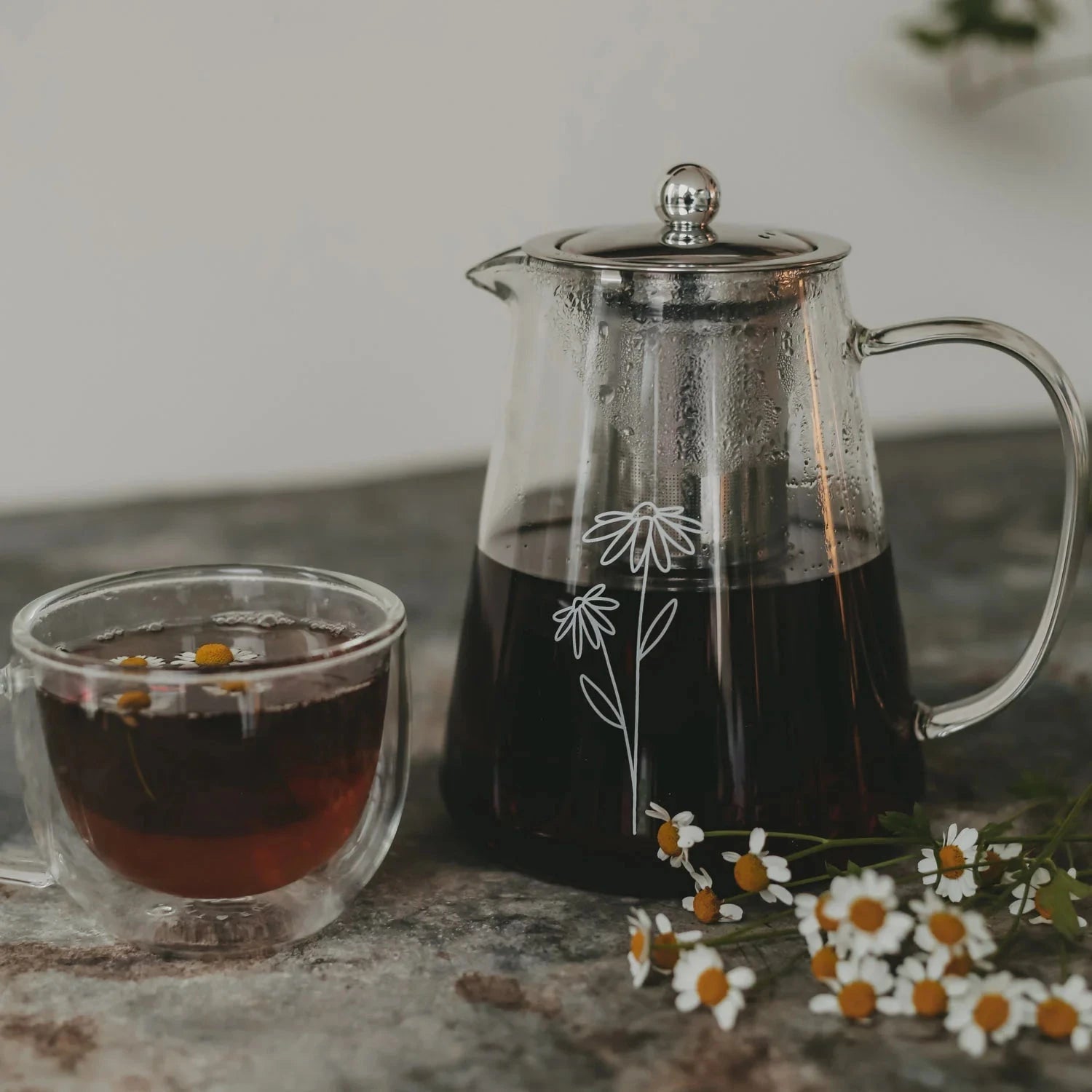 Teekanne "Daisy" aus Glas mit weißem Blumenaufdruck, gefüllt mit Tee, daneben liegen Margeriten und steht eine Tasse mit Tee.