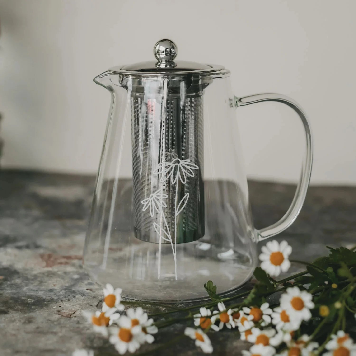 Teekanne "Daisy" aus Glas mit weißem Blumenaufdruck, daneben liegen Margeriten.