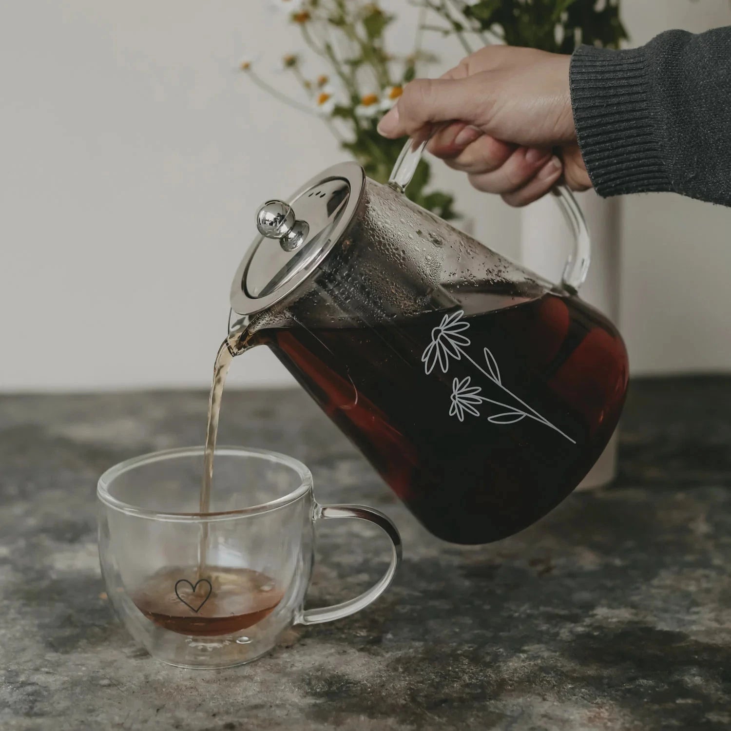 Teekanne "Daisy" aus Glas mit weißem Blumenaufdruck, füllt eine Tasse mit Tee.