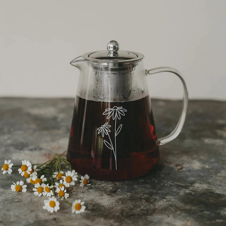 Teekanne "Daisy" aus Glas mit weißem Blumenaufdruck, gefüllt mit Tee, daneben liegen Margeriten.