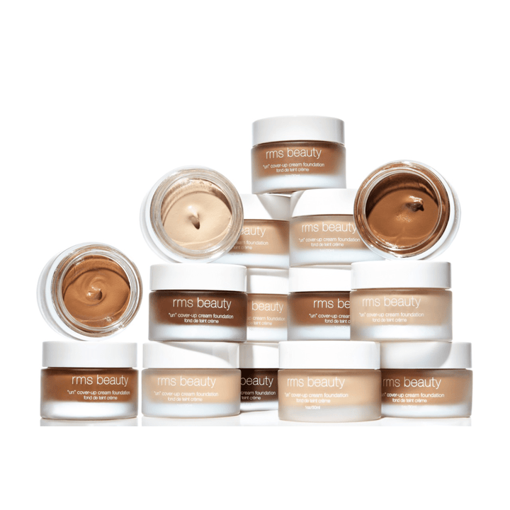 15 Glastiegel UnCover-up Cream Foundation, in verschiedenen Farben, davon 3 geöffnet, aufeinander gestapelt vor weißem Hintergrund.