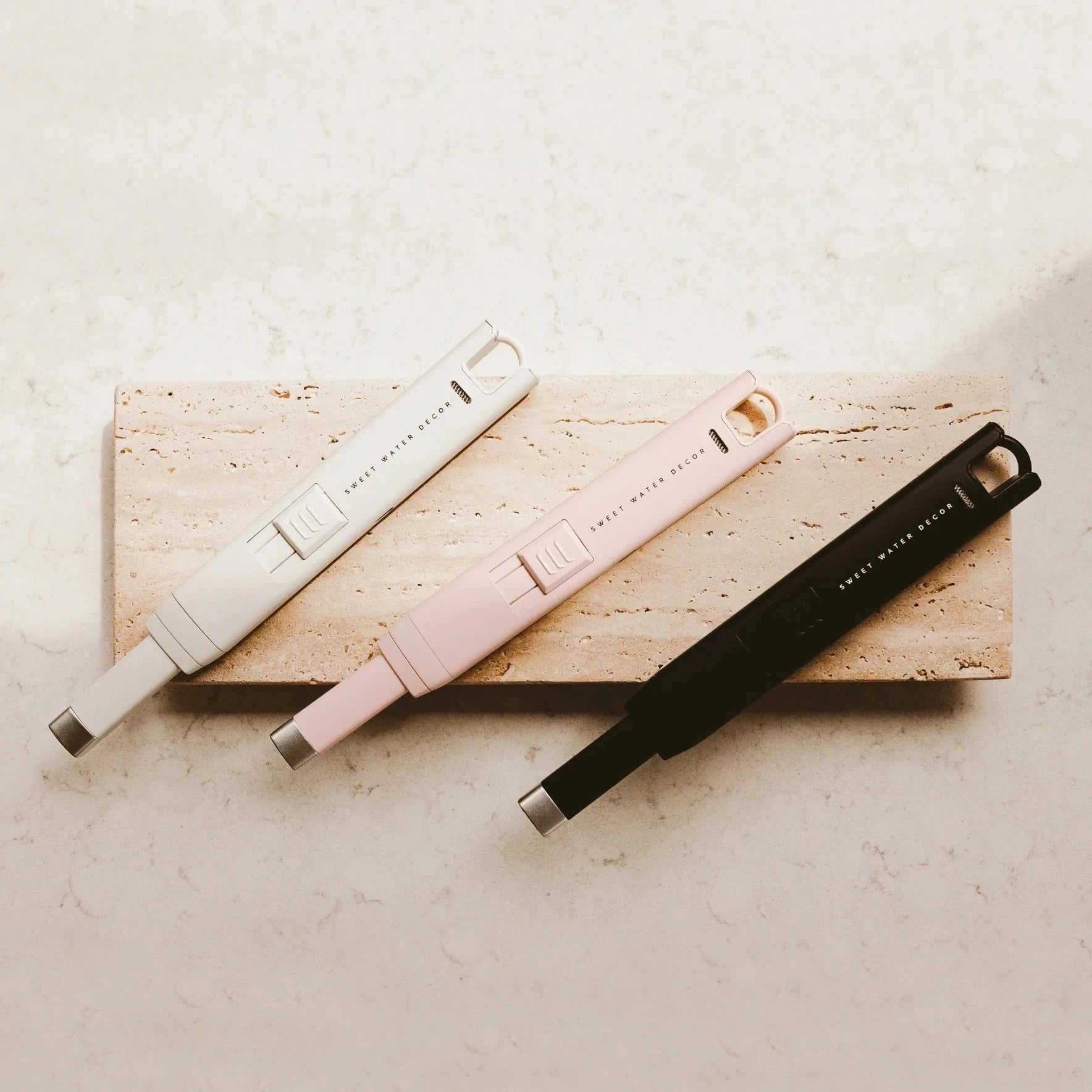 3 USB rechargable Feuerzeuge SWEET WATER DECOR in weiß, rosa und schwarz aus der Vogelperspektiveliegen auf einer Steinplatte.
