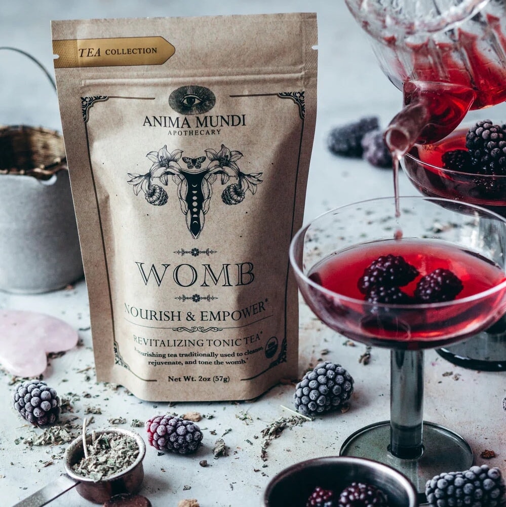 Anima Mundi Womb Tee Tüte steht zwischen Brombeeren und einem Glas mit roter Flüssigkeit.