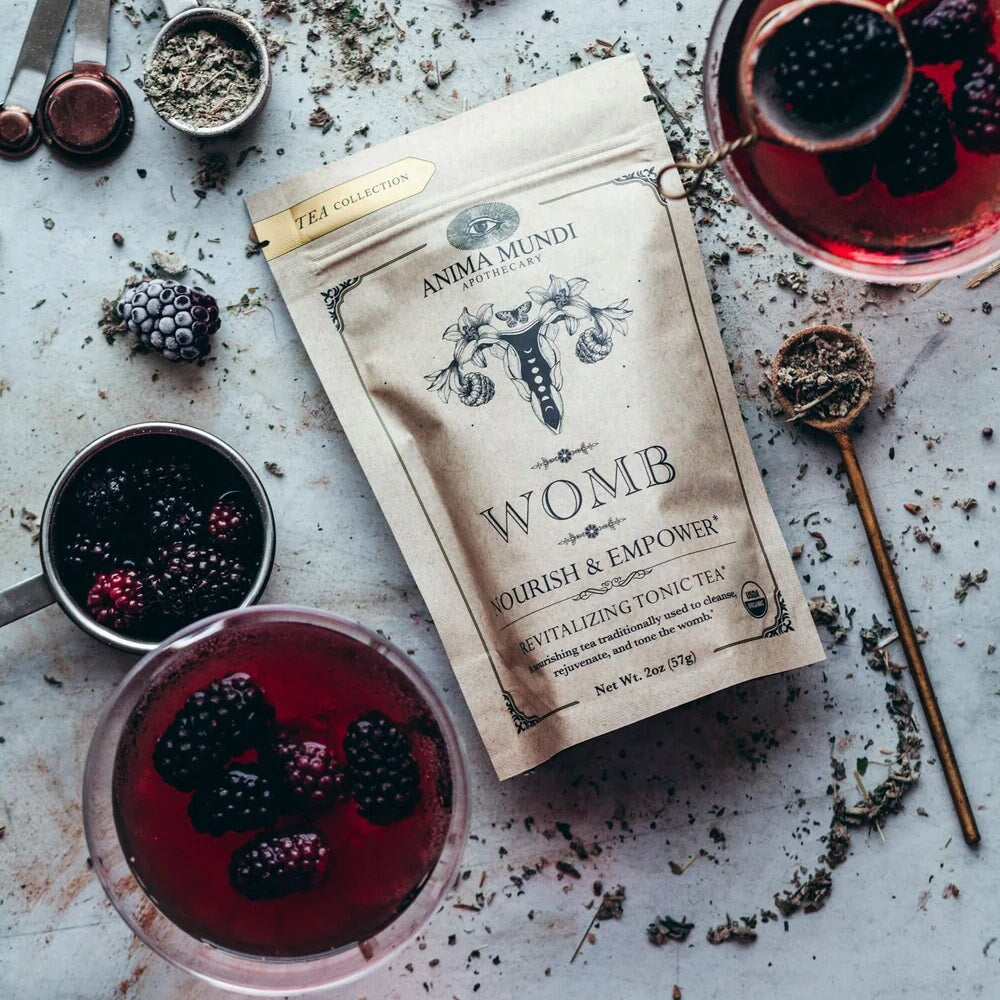 Anima Mundi Womb Tee Tüte aus der Vogelperspektive, liegt zwischen Gläsern mit roter Flüssigkeit und Brombeeren. North Glow