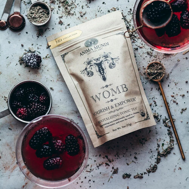Anima Mundi Womb Tee Tüte aus der Vogelperspektive, liegt zwischen Gläsern mit roter Flüssigkeit und Brombeeren.