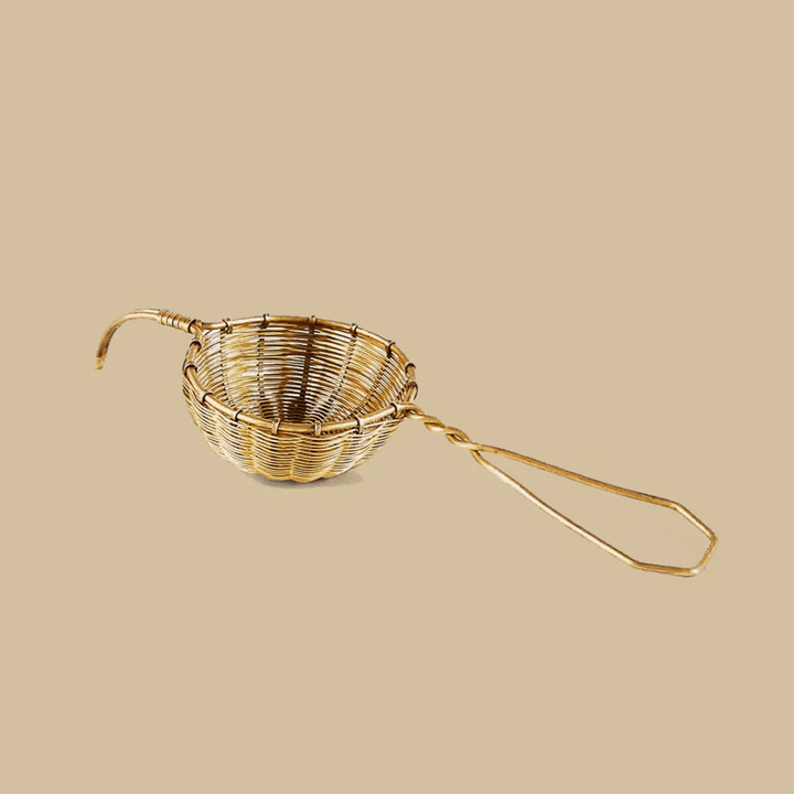 Woven Brass Small Tea Strainer, kleines Teesieb aus Messing vor cremefarbenem Hintergrund.