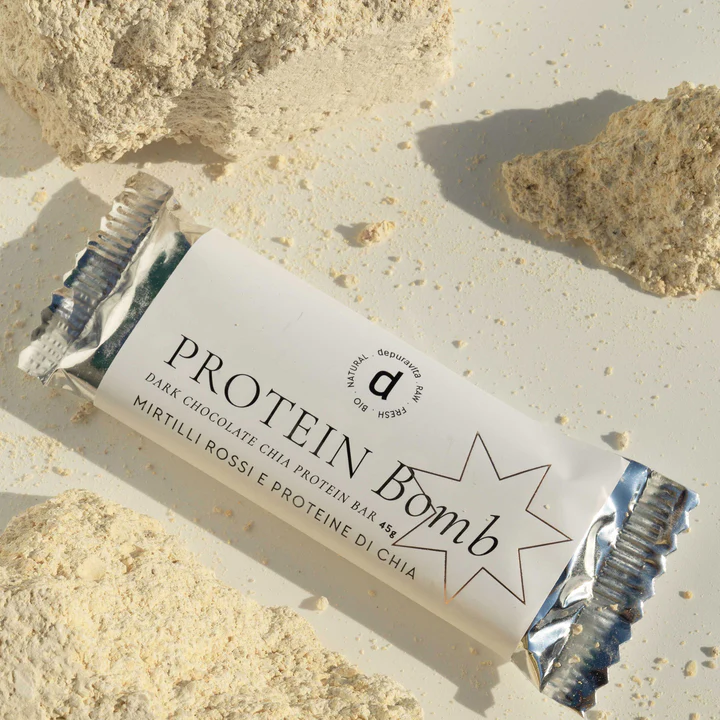 Depuravita Protein Bomb Riegel vor hellem sandigen Hintergrund