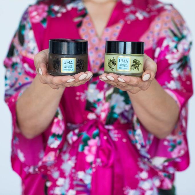 Im Hintergrund verschwommen der Oberkörper einer Frau bekleidet mit einem Kimono, hält in beiden Händen im Vordergrund UMA Absolute Anti Aging Body Scrub und Body Balm Schraubgläser in den Händen.