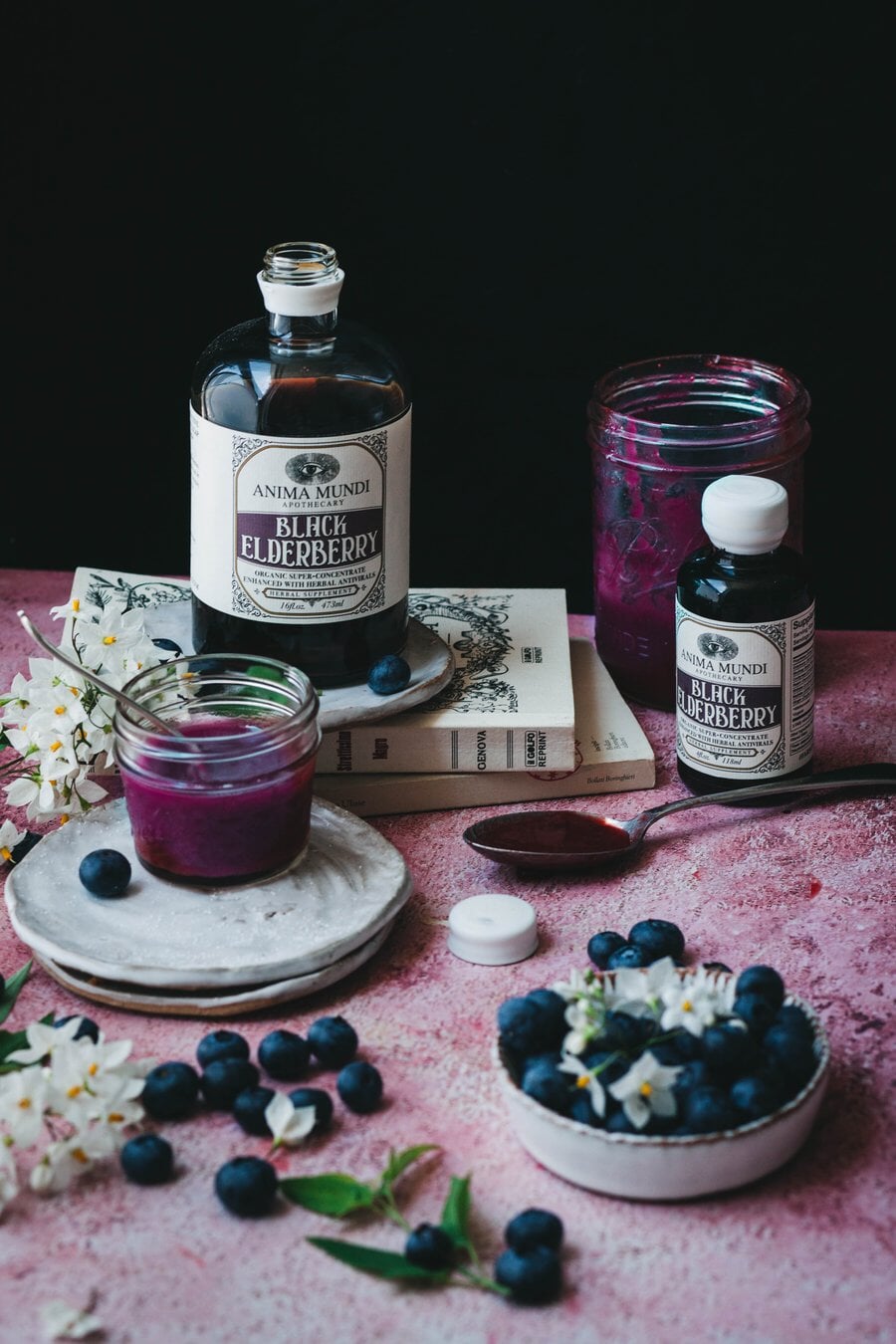 Anima Mundi Glasflasche Black Elderberry in 2 Größen stehen auf einem Tisch, dekoriert mit Holunderblüten und -beeren.