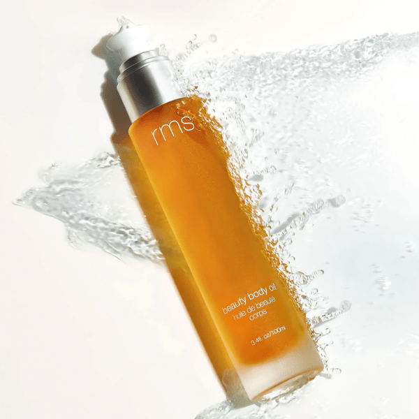 Flasche RMS Beauty Body Oil von einer kleinen Welle Wasser umspielt.