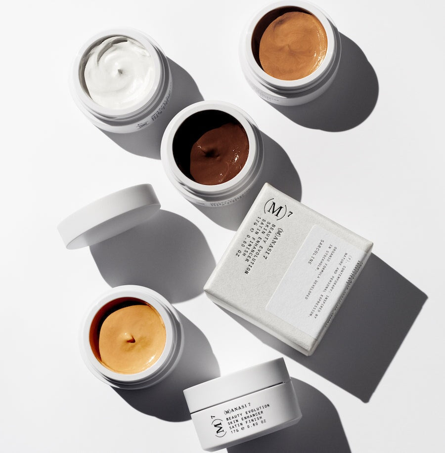 Verschiedene offene Tiegel der Manasi 7 Skin Enhancer stehen verstreut auf weißem Untergrund.