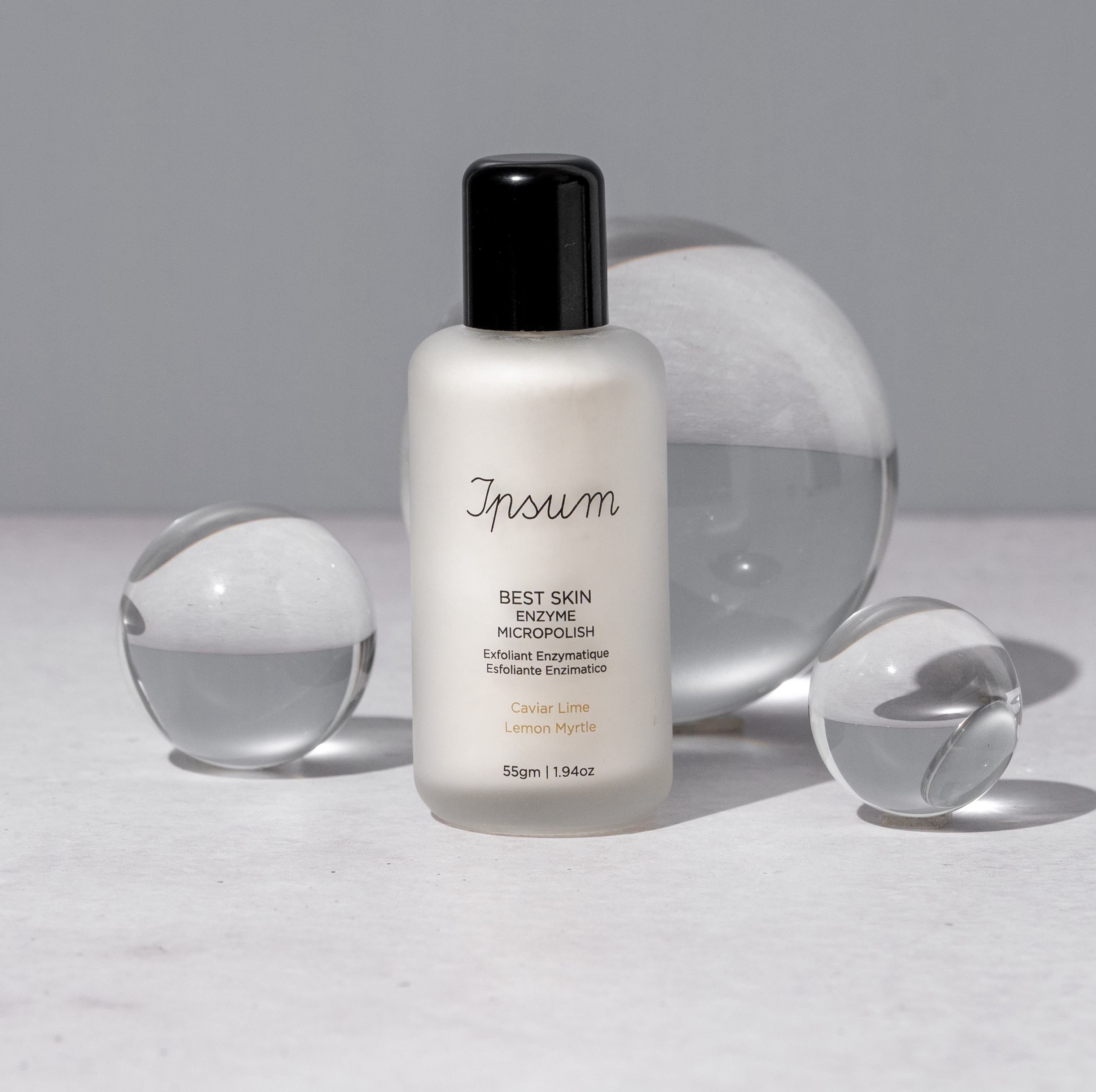 Ipsum Best Skin Enzyme MicroPolish Milchglasflasche mit schwarzem Deckel steht zwischen verschieden großen Glaskugeln.