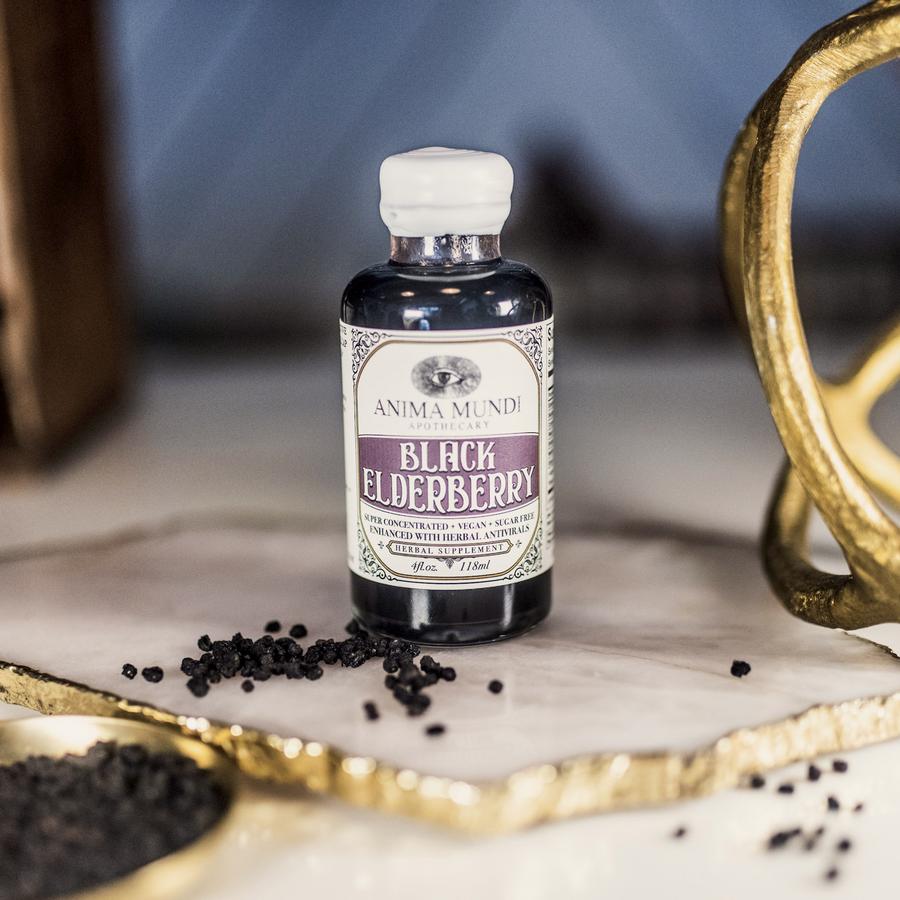 Anima Mundi Glasflasche Black Elderberry steht auf einer goldumrandeten Platte, davor getrocknete Holunderbeeren.