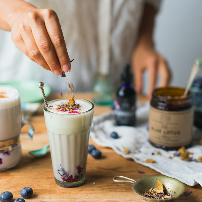 Eine Hand streut Blauen Lotus Teeblätter in ein Glas mit milchigem Tee, verschwommener Hintergrund mit Schraubverschlussglas Anima Mundi Blue Lotus Tee.