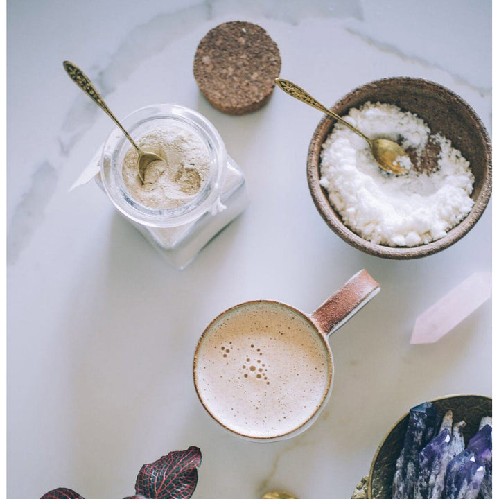 Aus der Vogelperspektive offenes Schraubglas mit hellem Pulver, Mörser mit Kokosnussraspeln und eine Tasse mit cremigen Getränk.