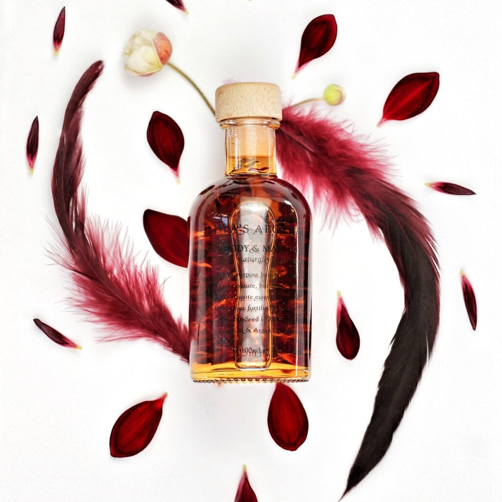 Lola´s Apothecary Delicate Romance Body & Massage Oil Flakon im Hintergrund Federn und Rosenblättern