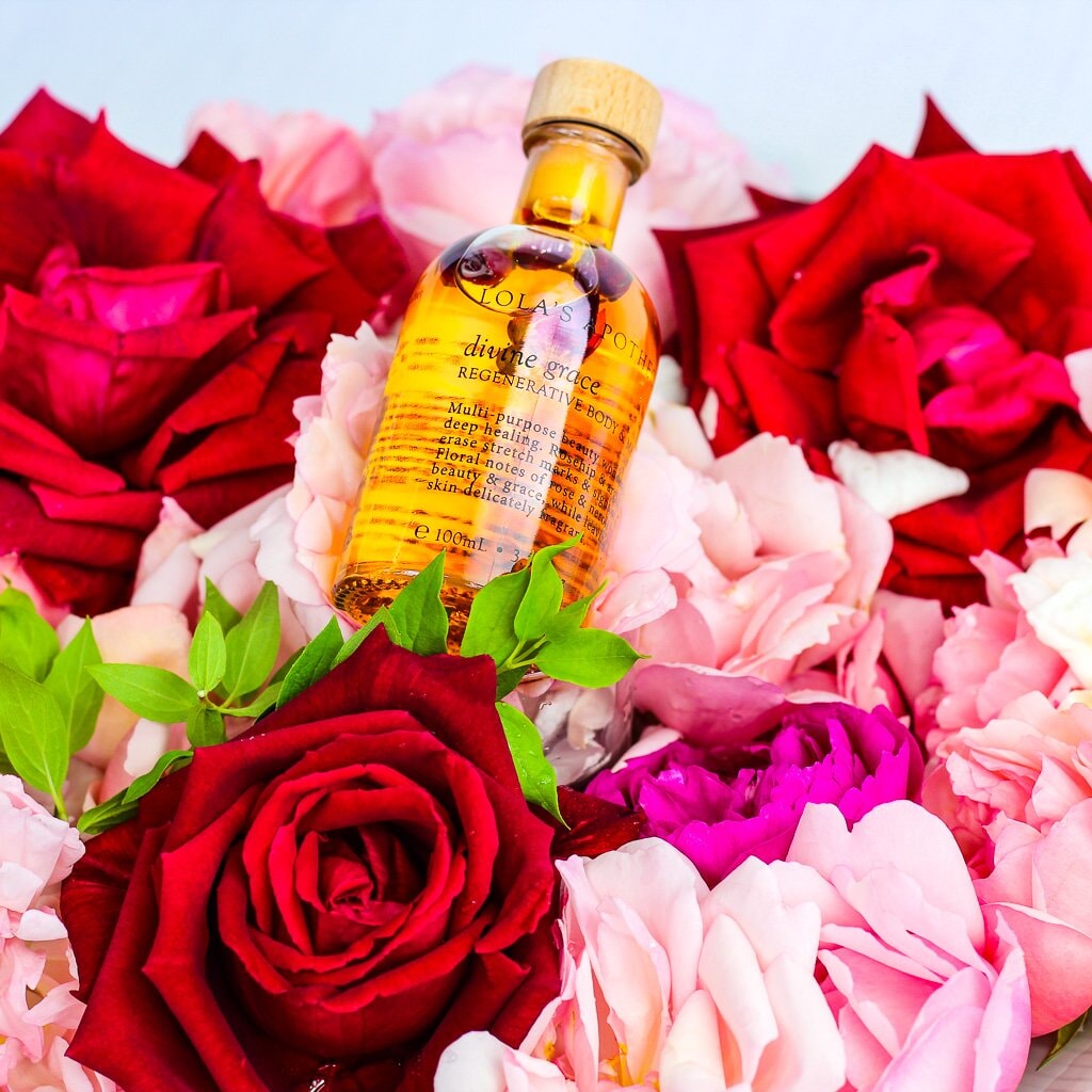 Glasflasche Lola´s Apothecary Body & Massageöl mit orangerotem Inhalt, liegend auf Rosen.