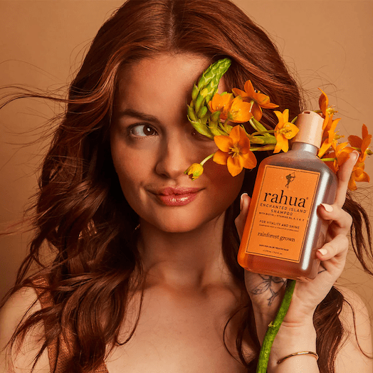 Frau mit roten Haaren vor orangefarbenem Hintergurnd hält Rahua Enchanted Island Shampoo Flasche. North Glow