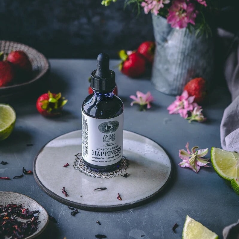 Anima Mundi Glaspipettenflasche Happiness steht auf einer Steinplatte zwischen Erdbeere, Limettenscheiben, Blütenblättern und getrockneten Kräutern.