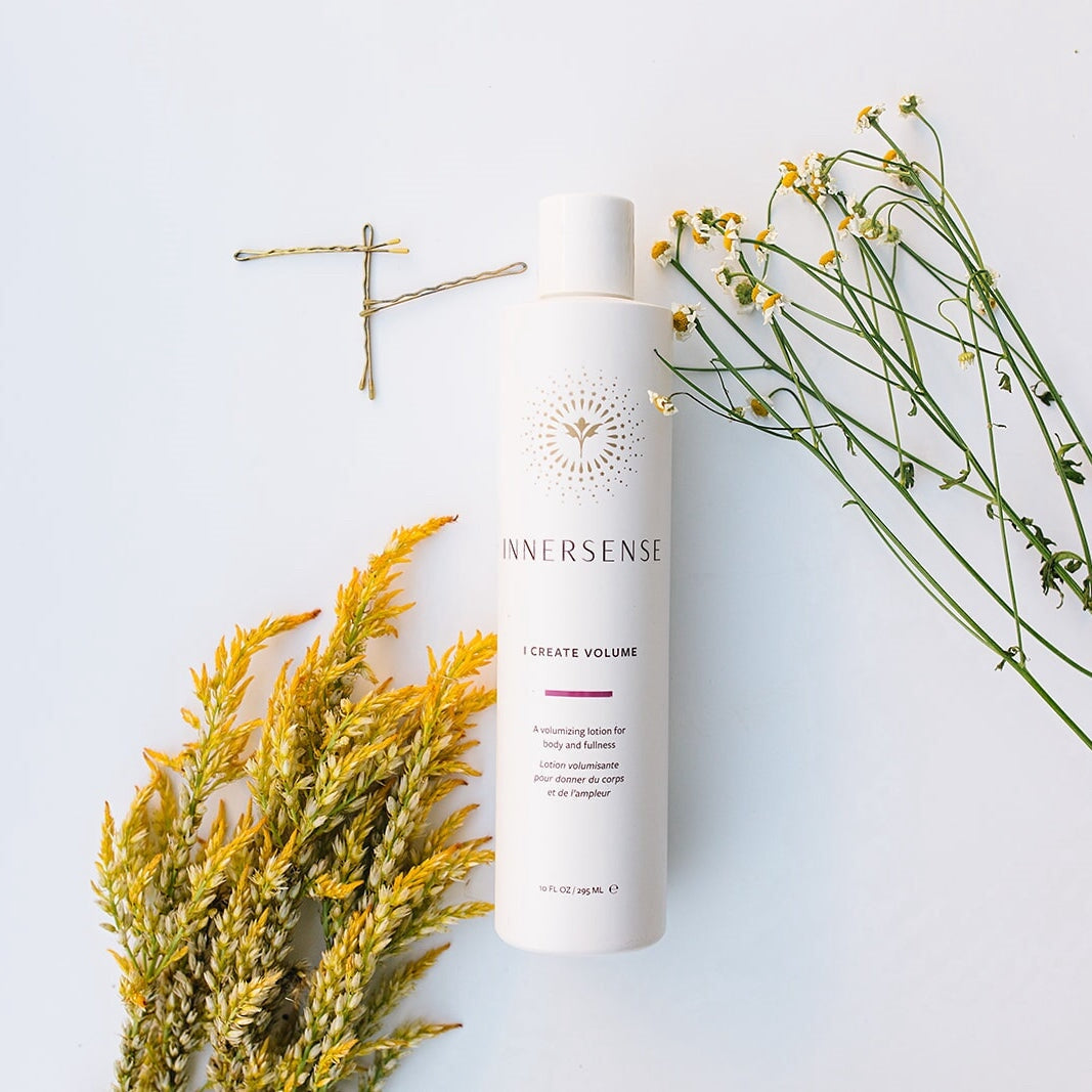 Eine weiße Innersense I Create Volume Flasche umringt von Haarnadeln und getrockneten Blüten auf weißem Untergrund.