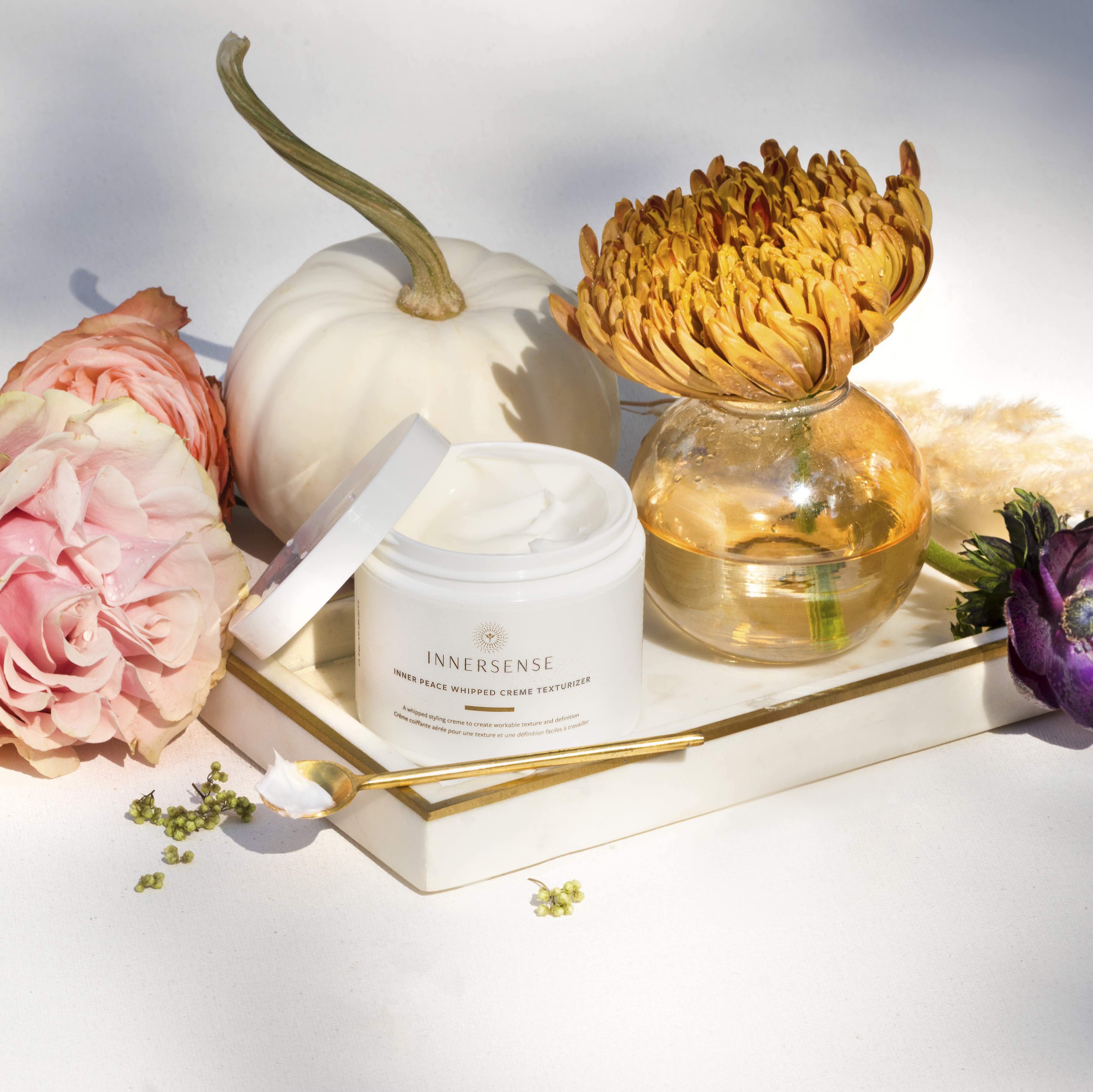 Offener Tiegel Innersense Inner Peace Whipped Creme Texturizer Tiegel steht zwischen weißem Kürbis, verschiedenfarbigen Blumen und einem Messinglöffel.